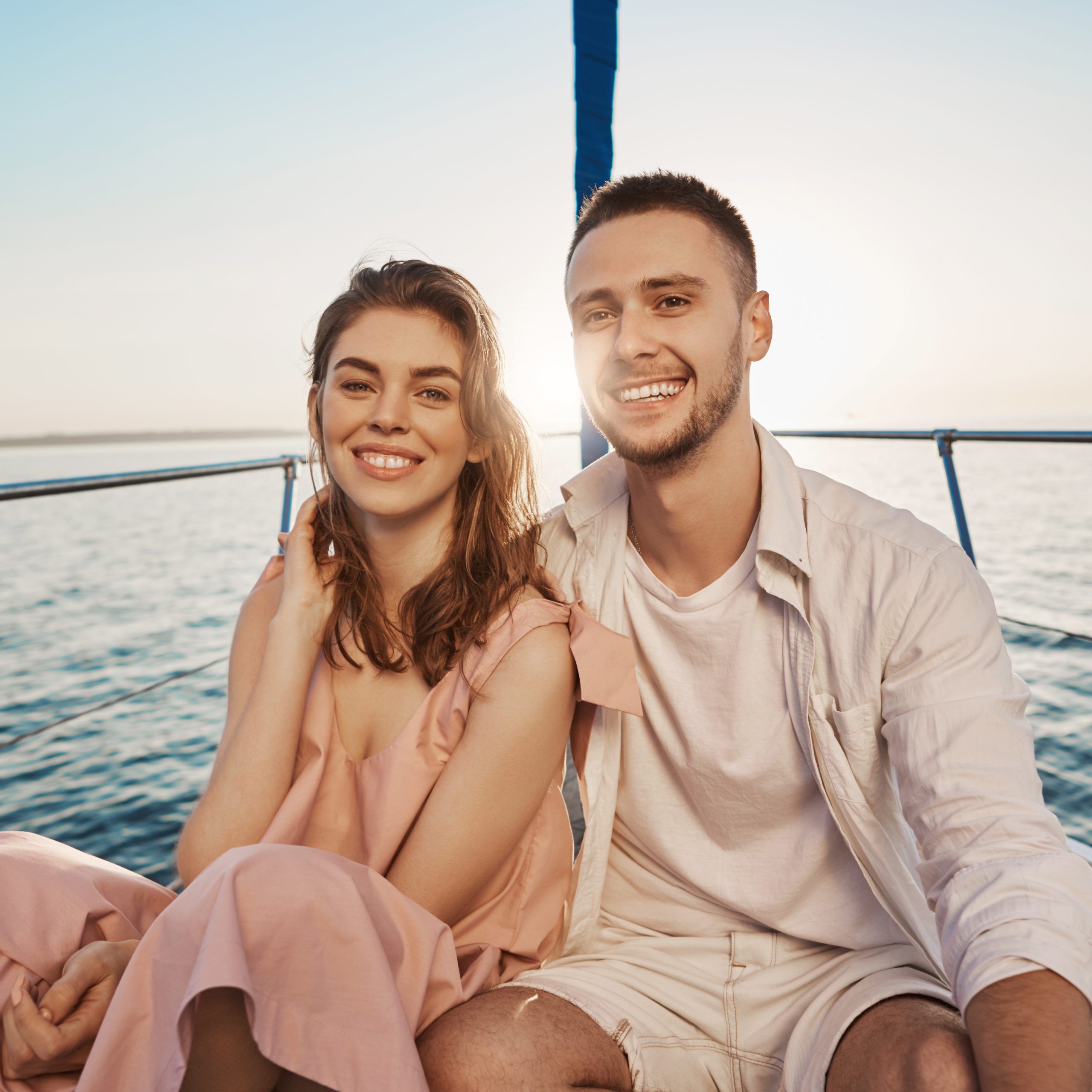 Young european romantic couple smiling at camera while sitting at bow of boat, hugging, enjoying their holidays. Two close friends recently became something more to each other